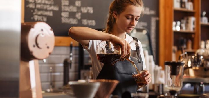 A young female barista in a black apron and white T-shirt prepares a pour-over coffee in a coffee shop.