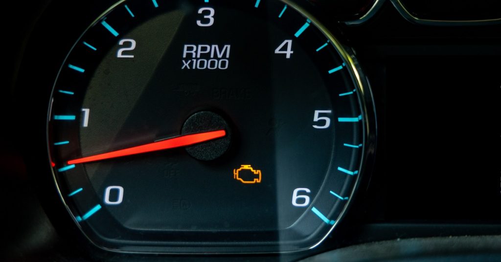 A close-up of a black vehicle dashboard with a circular tachometer showing an orange check engine light.
