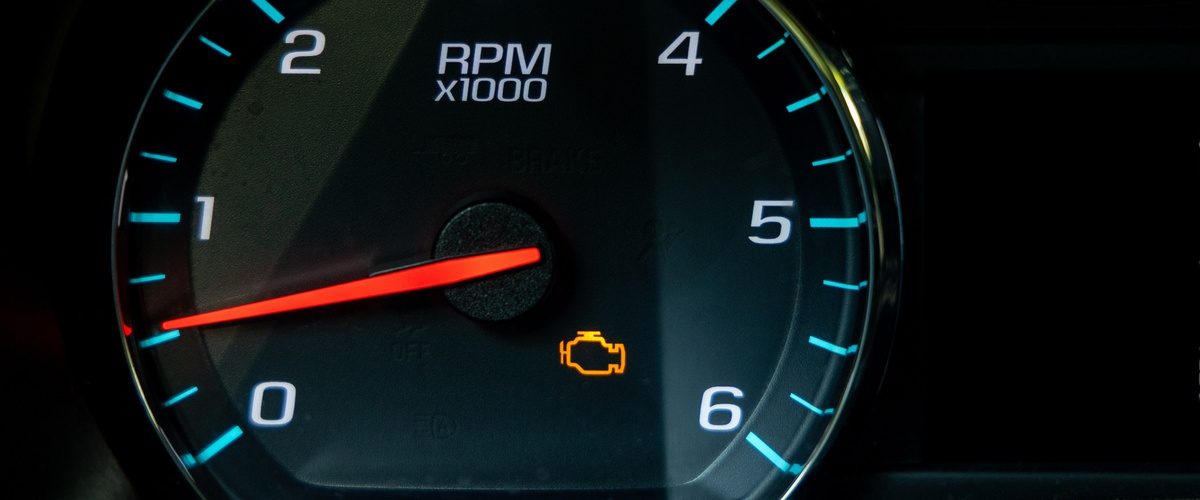A close-up of a black vehicle dashboard with a circular tachometer showing an orange check engine light.