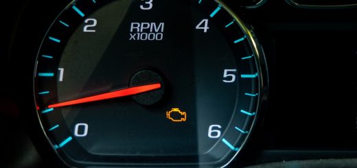 A close-up of a black vehicle dashboard with a circular tachometer showing an orange check engine light.