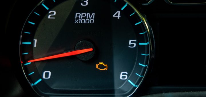 A close-up of a black vehicle dashboard with a circular tachometer showing an orange check engine light.