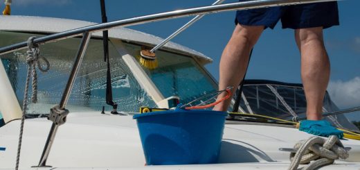 A close-up of the bottom half of a man wearing navy shorts washing a white boat with a brush and a pressure water system.