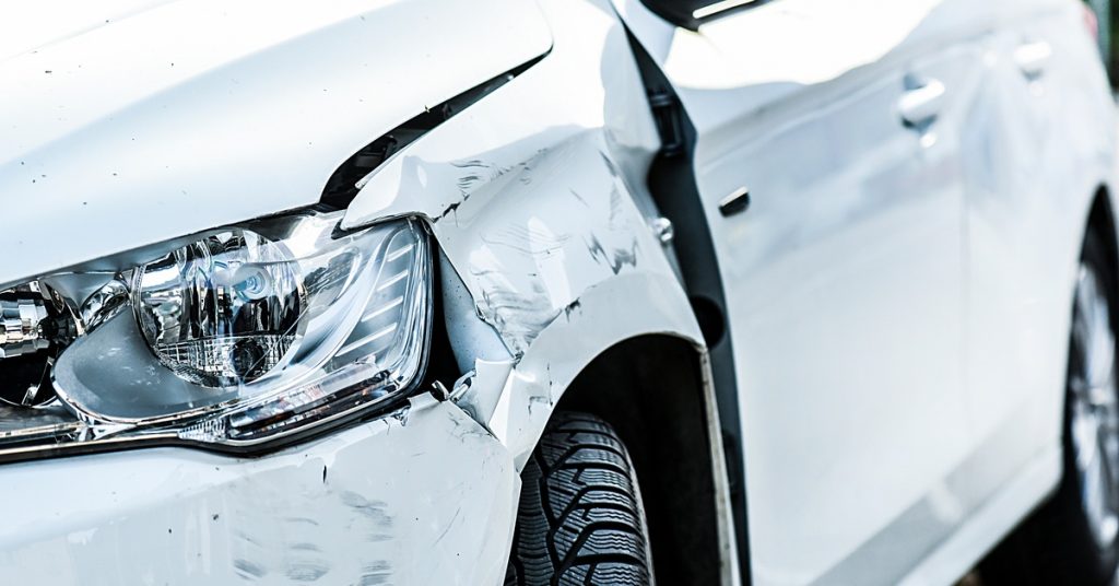 The front end of a white car with damage from an accident. The side panel next to the headlight is dented and scuffed.