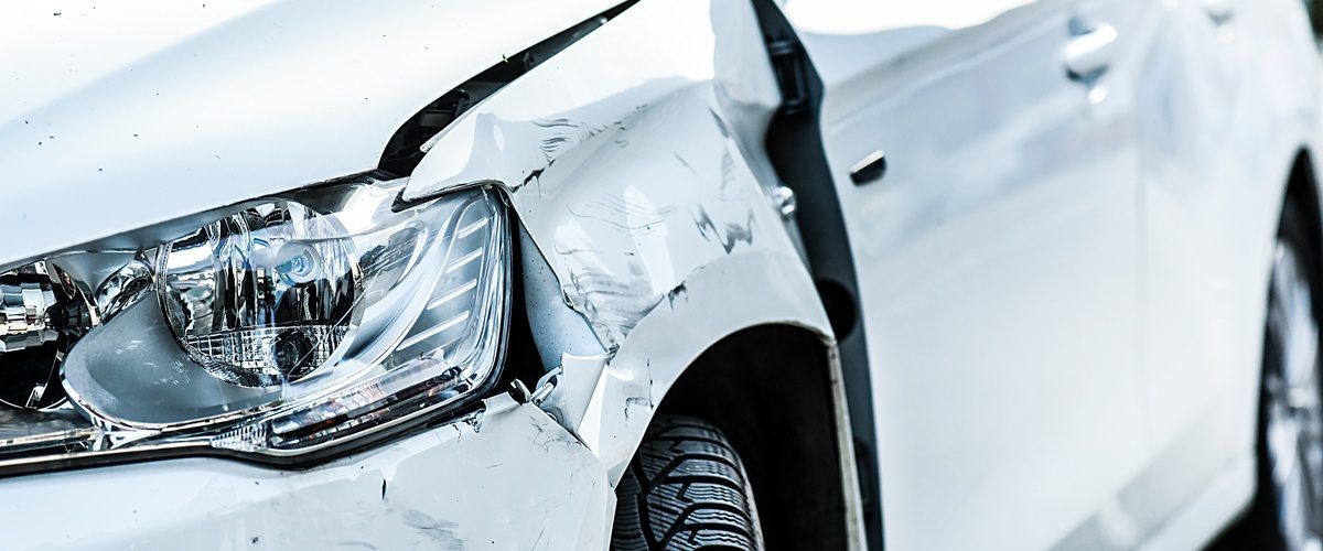 The front end of a white car with damage from an accident. The side panel next to the headlight is dented and scuffed.