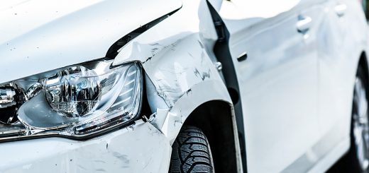 The front end of a white car with damage from an accident. The side panel next to the headlight is dented and scuffed.