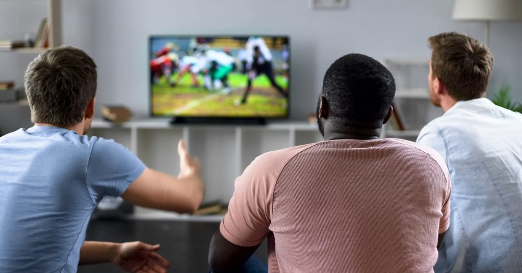Rear view of three men, watching sports together on a TV screen. One holds up his hand in frustration.
