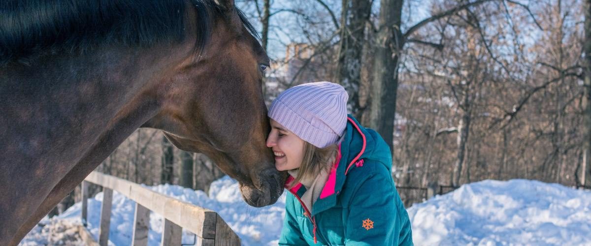A smiling woman wearing a teal winter coat and white stocking cap is leaning on wooden fence and pressing her cheek against the face of a brown horse.