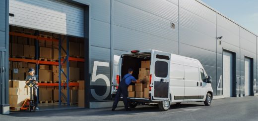 Two people are loading up a white commercial delivery truck with cardboard boxes. It is parked next to a warehouse.