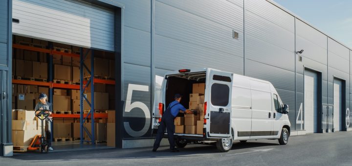 Two people are loading up a white commercial delivery truck with cardboard boxes. It is parked next to a warehouse.