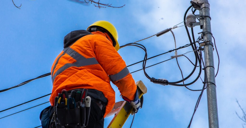 A fiber optics professional wears a high-vis orange coat and a yellow hardhat as they stand near the electrical pole.