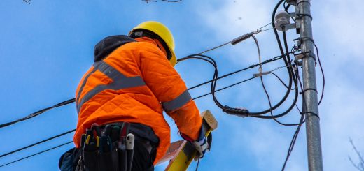 A fiber optics professional wears a high-vis orange coat and a yellow hardhat as they stand near the electrical pole.