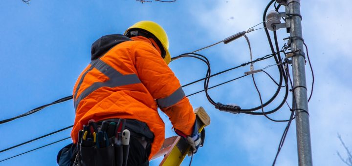 A fiber optics professional wears a high-vis orange coat and a yellow hardhat as they stand near the electrical pole.