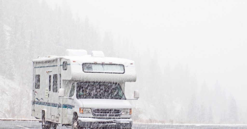 Snow is falling heavily on an RV parked in an empty parking lot. A hill of pine trees is obscured by the snowfall behind it.