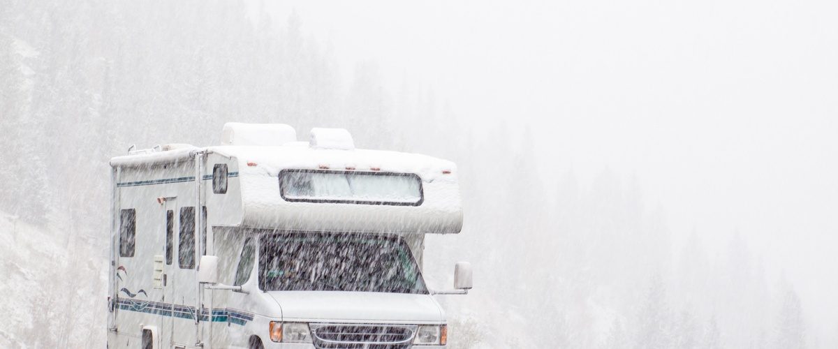 Snow is falling heavily on an RV parked in an empty parking lot. A hill of pine trees is obscured by the snowfall behind it.