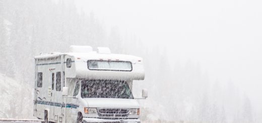 Snow is falling heavily on an RV parked in an empty parking lot. A hill of pine trees is obscured by the snowfall behind it.