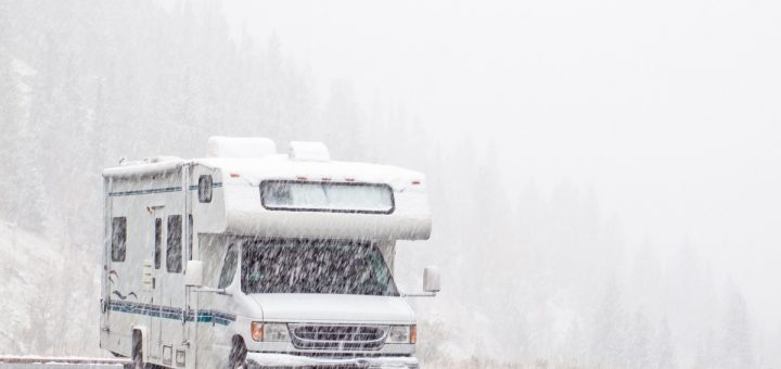 Snow is falling heavily on an RV parked in an empty parking lot. A hill of pine trees is obscured by the snowfall behind it.