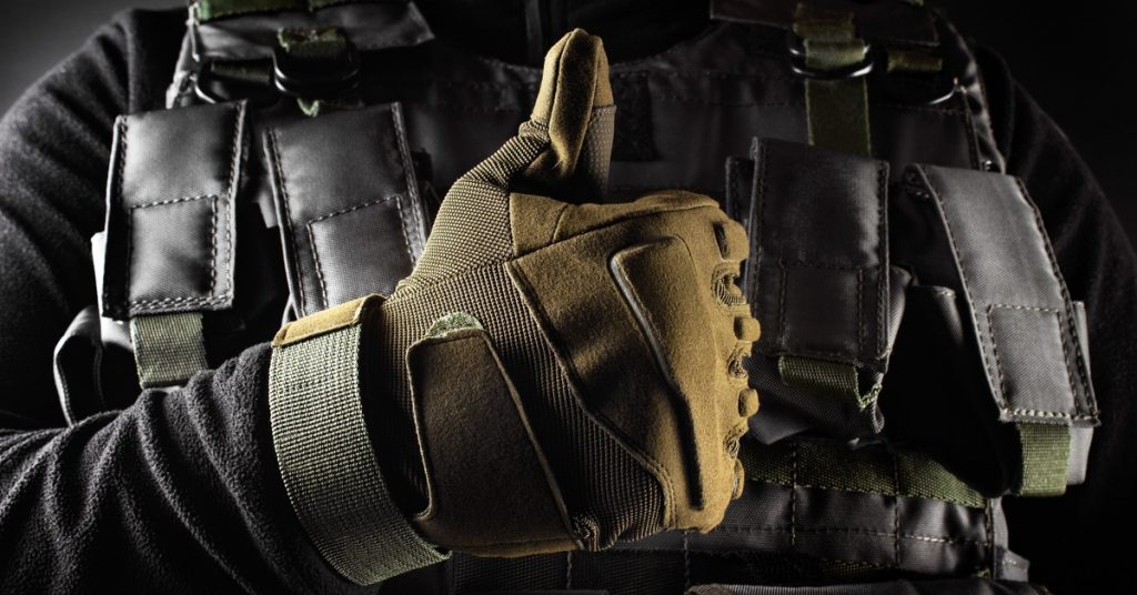 A close-up of a person wearing military-grade tactical gear, such as a bulletproof vest and safety gloves, with a thumbs-up.