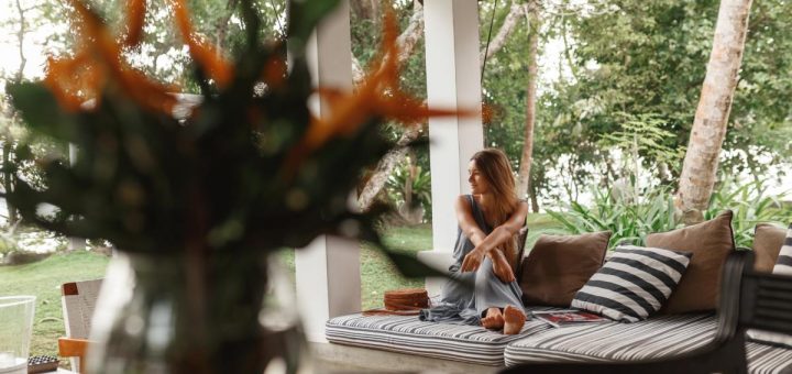 A woman with long hair is sitting on a chair in a yard. There is a plant in the foreground.