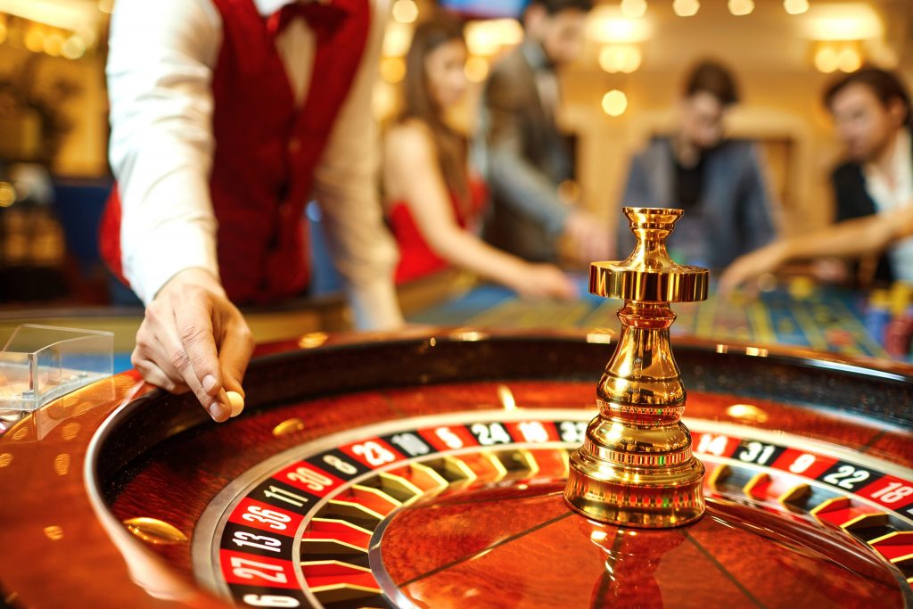 The hand of a casino dealer holding a small white ball on top of a roulette wheel with people in the background.