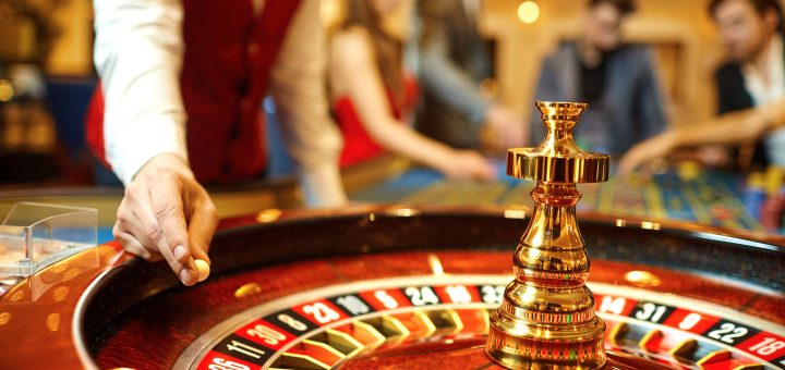 The hand of a casino dealer holding a small white ball on top of a roulette wheel with people in the background.
