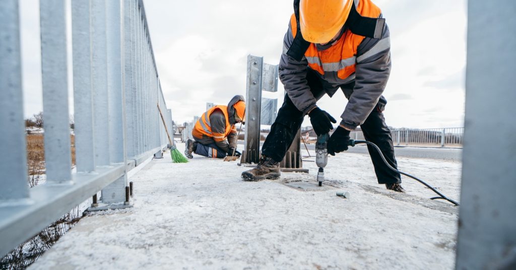Two men are wearing orange high-visibility vests. They are holding tools and working on an unfinished structure.