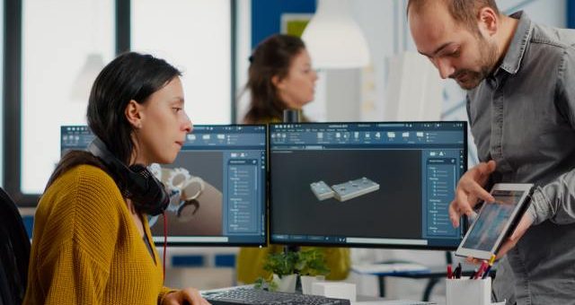 A man showing a tablet to a woman behind a desk with two large computer monitors displaying a product design render.