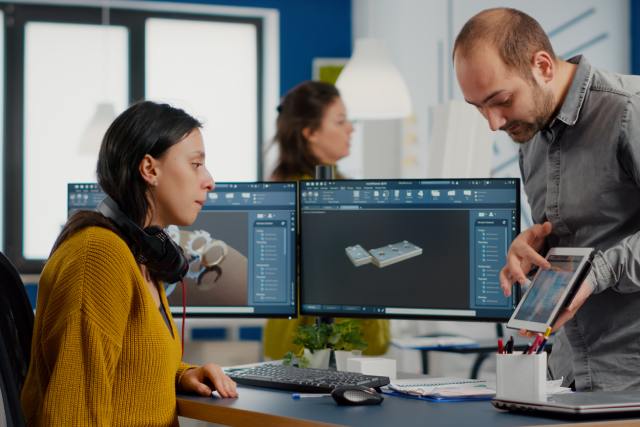 A man showing a tablet to a woman behind a desk with two large computer monitors displaying a product design render.