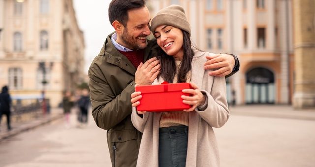 A man stands with a woman in the middle of a city. Both are smiling, and she holds a red box that looks like a present.
