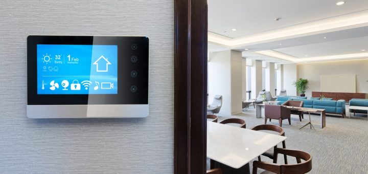 A smart thermostat with a digital screen installed on a wall in an office with tables, chairs, and couches.