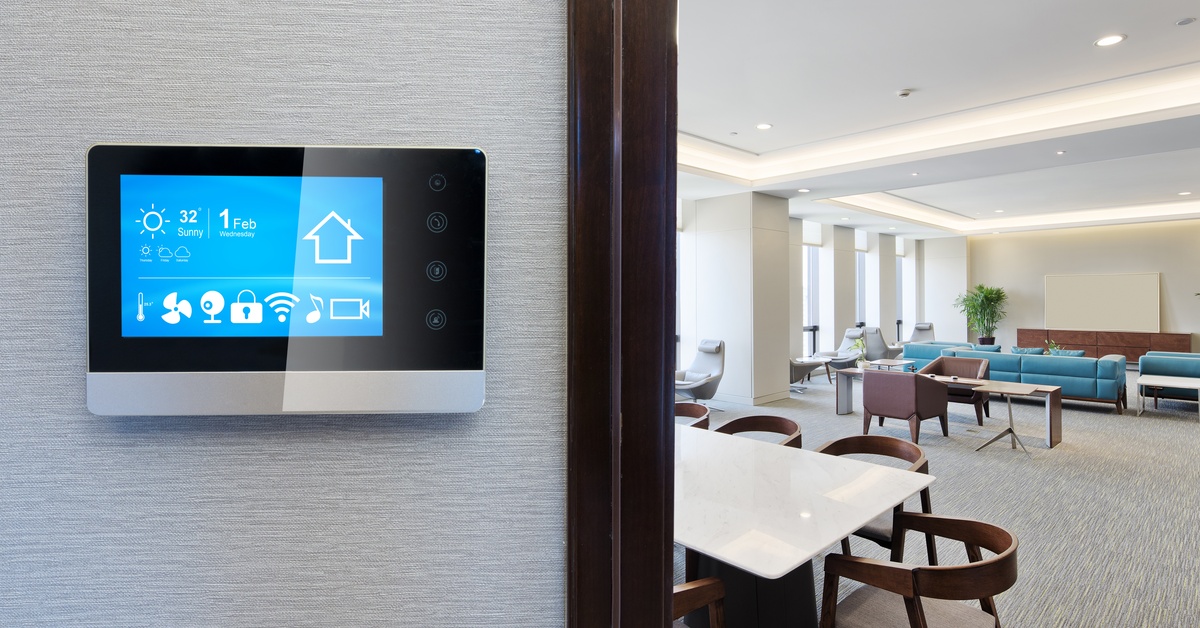 A smart thermostat with a digital screen installed on a wall in an office with tables, chairs, and couches.