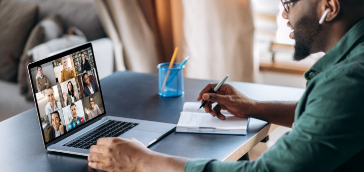 A man is sitting at a desk with his laptop open. On the screen, there are other people in a video call.