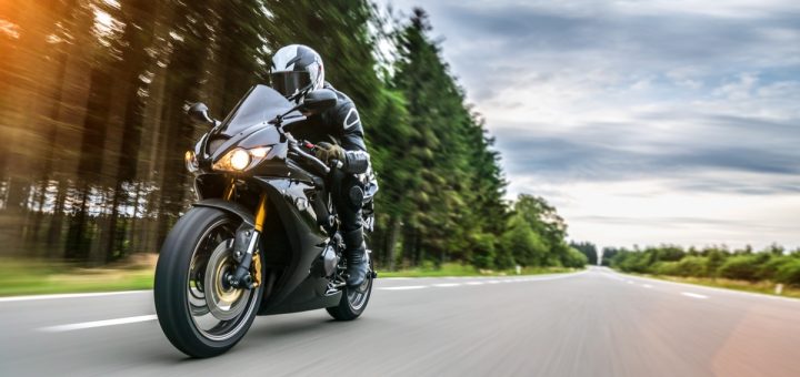 A person rides on a motorcycle along an empty road. There are trees behind them on the side of the road.