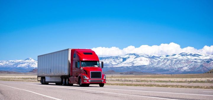 A semi-truck with a bright red cab driving on an open road with snow-capped mountains in the background.