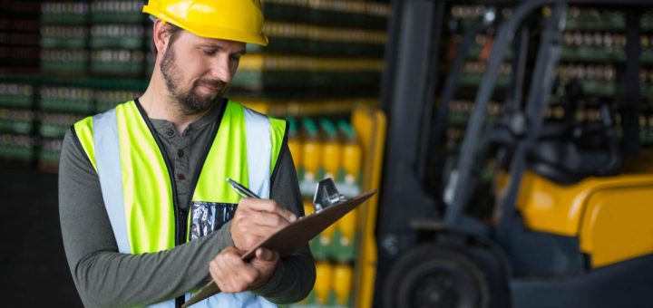 A man in a yellow hard hat and green safety vest writing on a clipboard with a pen with a forklift in the background.