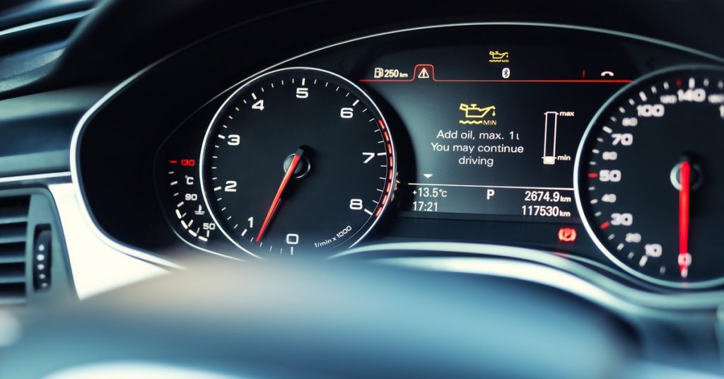 A car dashboard with analog speed and RPM gauges. The center display shows an engine oil warning and driving stats.