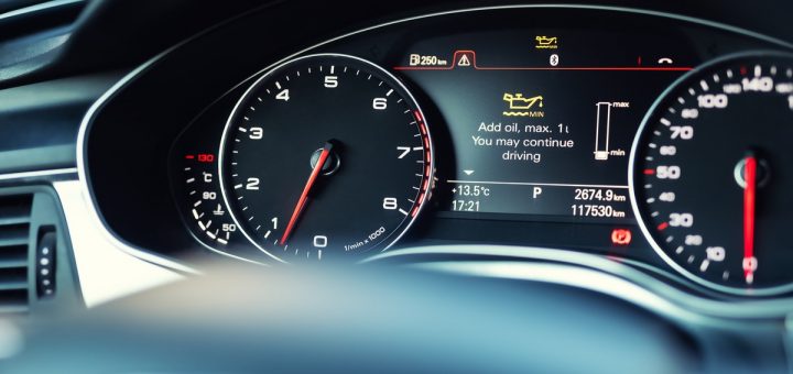 A car dashboard with analog speed and RPM gauges. The center display shows an engine oil warning and driving stats.