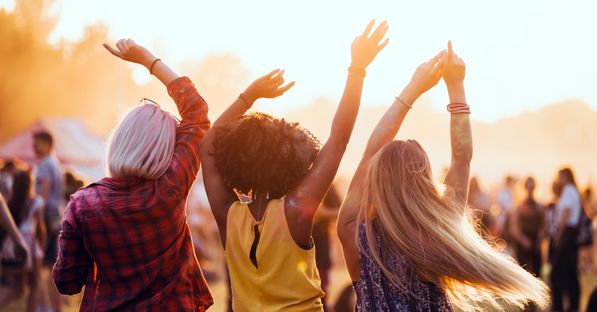 Three friends with raised arms enjoy a summer music festival at sunset with a crowd in the background.
