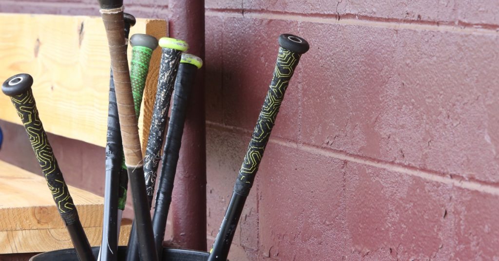 Seven black baseball bats upside down in a bucket, locked in a dugout with a red wall and wooden bench, during the day.