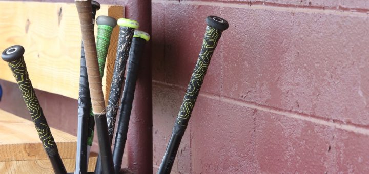 Seven black baseball bats upside down in a bucket, locked in a dugout with a red wall and wooden bench, during the day.
