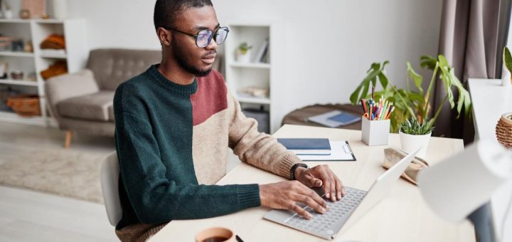 A man working from home. He sits at a desk in his living room with a coffee, a laptop, and other desk accessories.