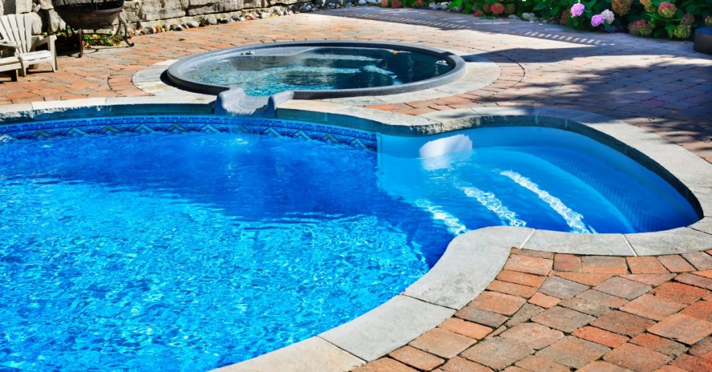 A close-up of a pool with four steps, a concrete edge, a brick path surrounding it and a small hot tub on the side.