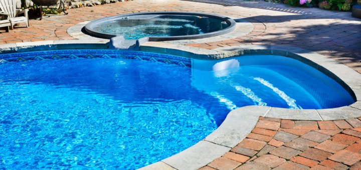 A close-up of a pool with four steps, a concrete edge, a brick path surrounding it and a small hot tub on the side.