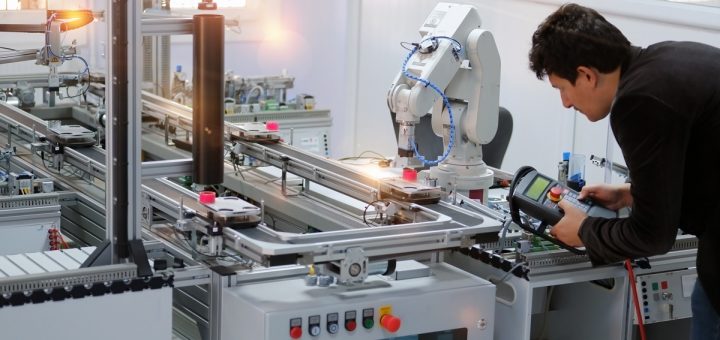 A man holding a large control with multiple buttons next to a large metal robotic arm attached to an industrial machine.
