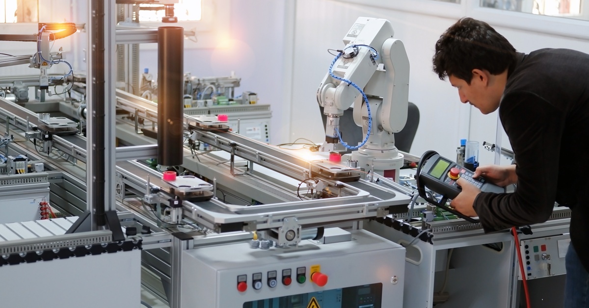 A man holding a large control with multiple buttons next to a large metal robotic arm attached to an industrial machine.