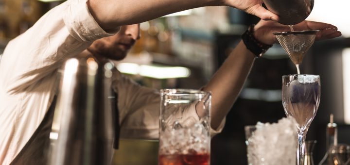 A young, bristled barman is making a cocktail while pouring it from the shaker into a strainer into a glass.