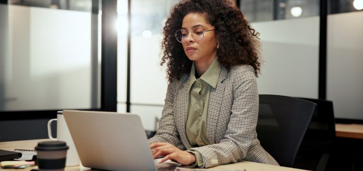 A businesswoman sits in her office and looks at her laptop. She wears glasses and has a coffee mug next to her.