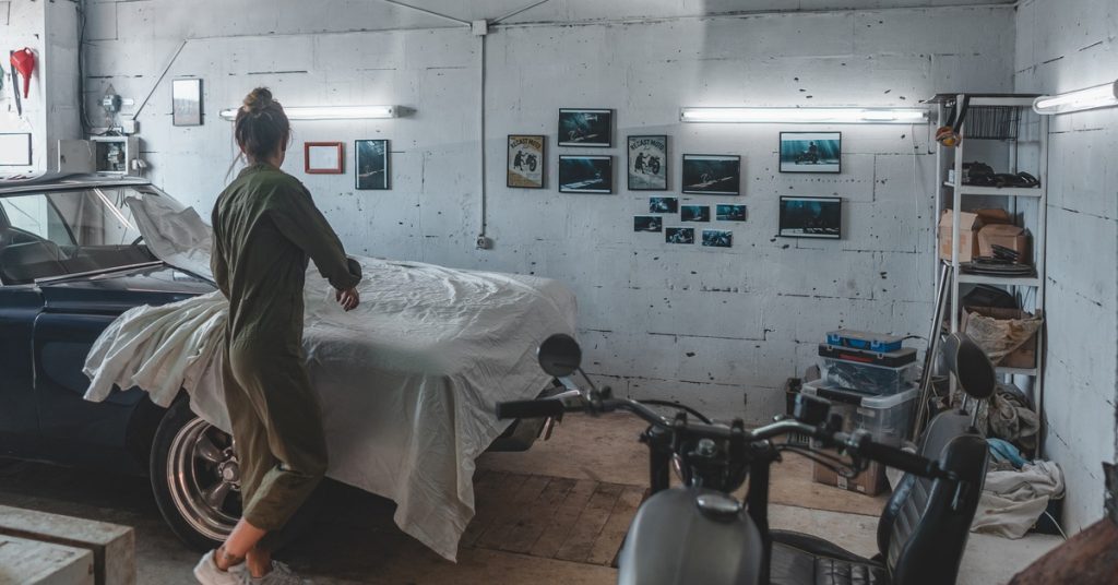 A woman in a green jumpsuit and tennis shoes in an old garage removing the cover of a vintage blue car.