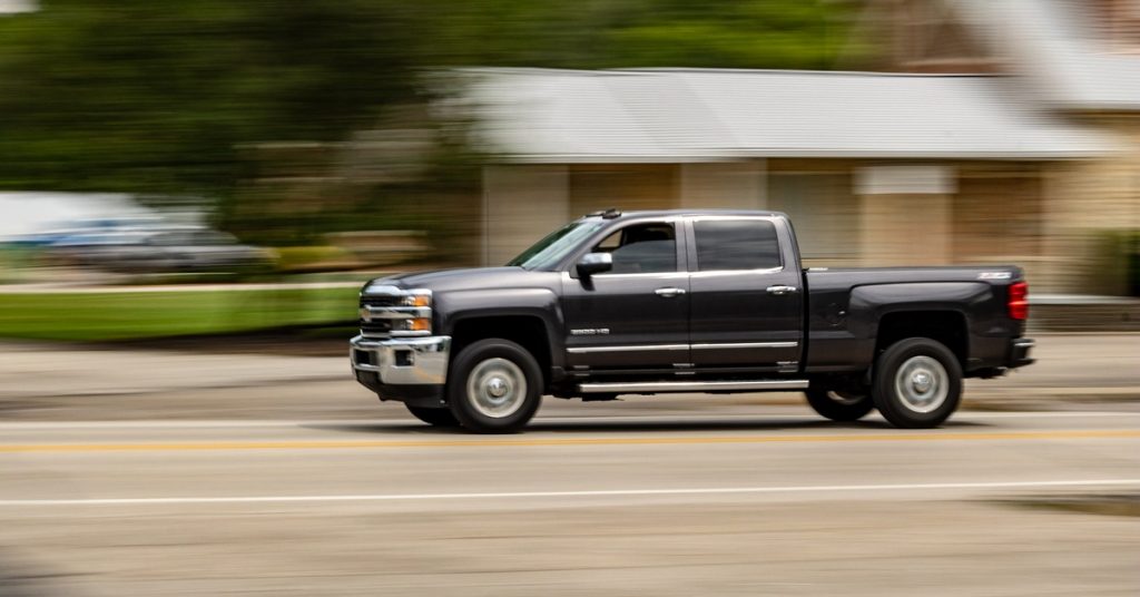A black four-door pickup truck is driving fast down the street past trees and commercial buildings.