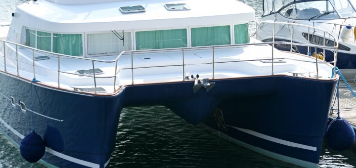 A white and blue catamaran with twin hulls floats in a marina, featuring a white deck, cabin, and metal railings.