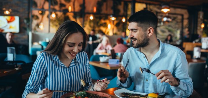 Two people, a man and a woman, are sitting at a table with eating utensils in their hands.
